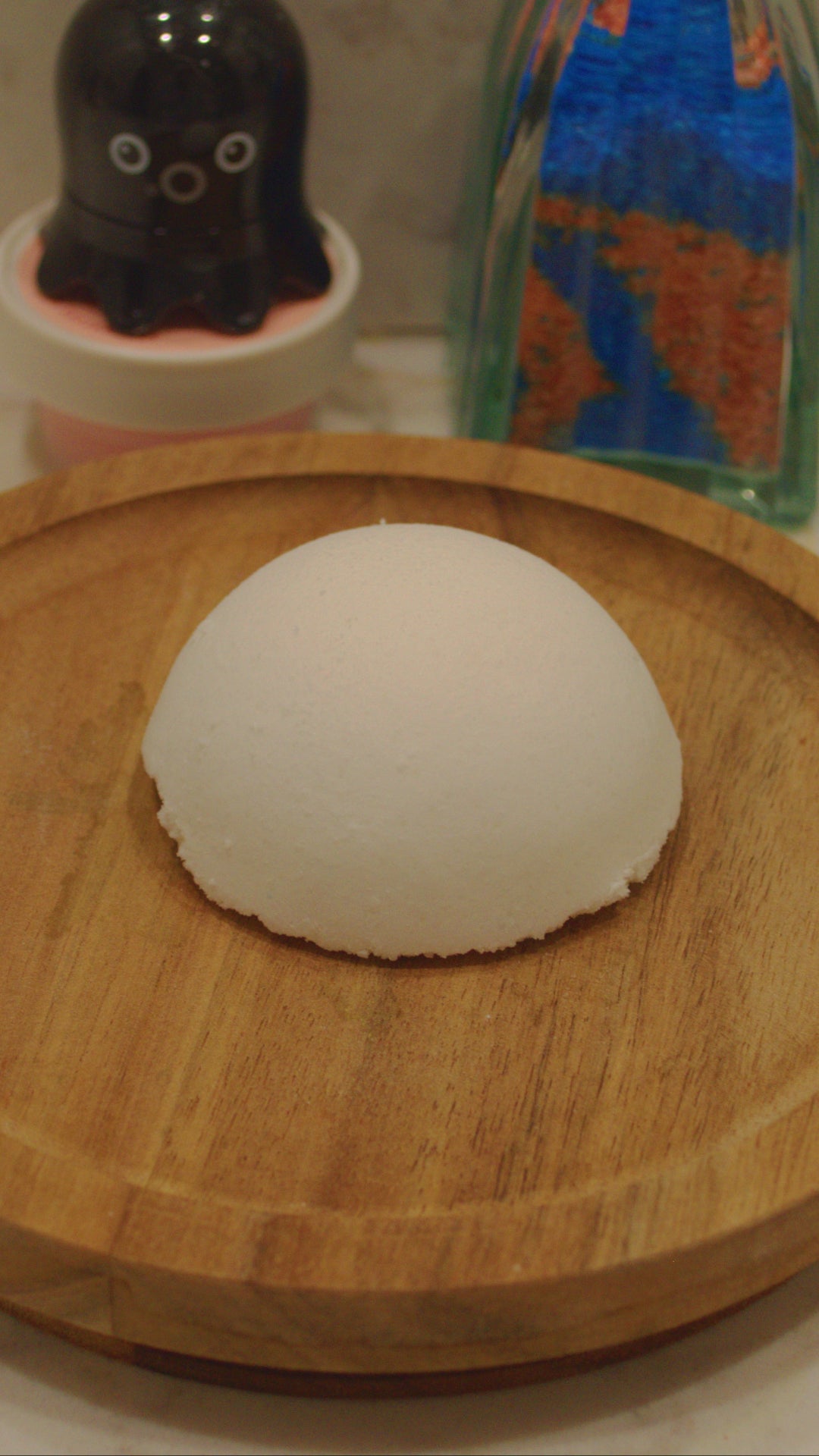 White bath bomb on a wooden tray with a black octopus-shaped bottle and a glass vase with colorful bubble bath powder in the background.