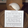 White bath bomb in a brown paper bag with an ingredients label on a wooden surface.